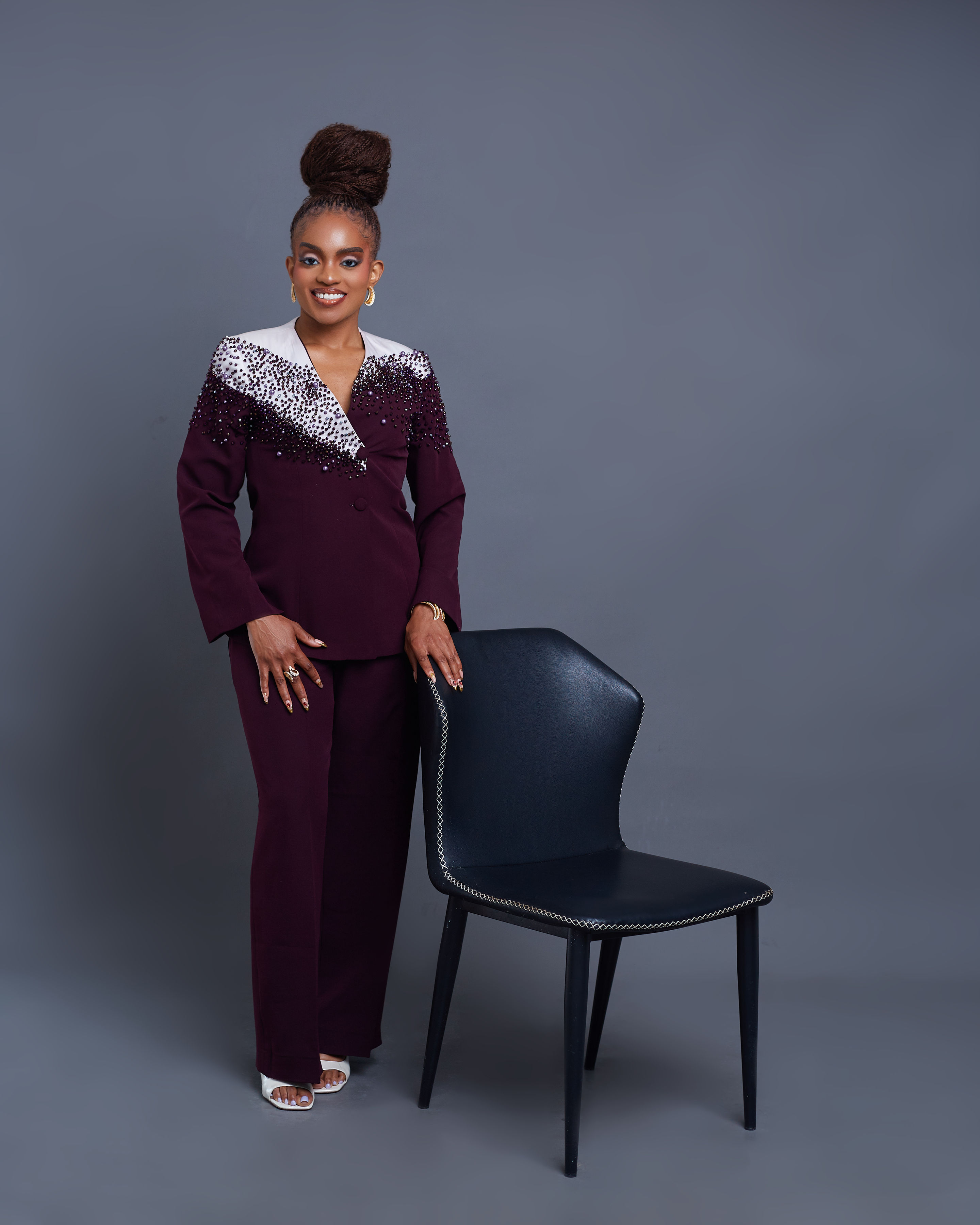 Full-length view of the Chikamso Suit — burgundy beaded jacket and matching wide-leg trousers, worn by founder Adaobi Onyekaba beside a black leather chair against a grey studio backdrop.