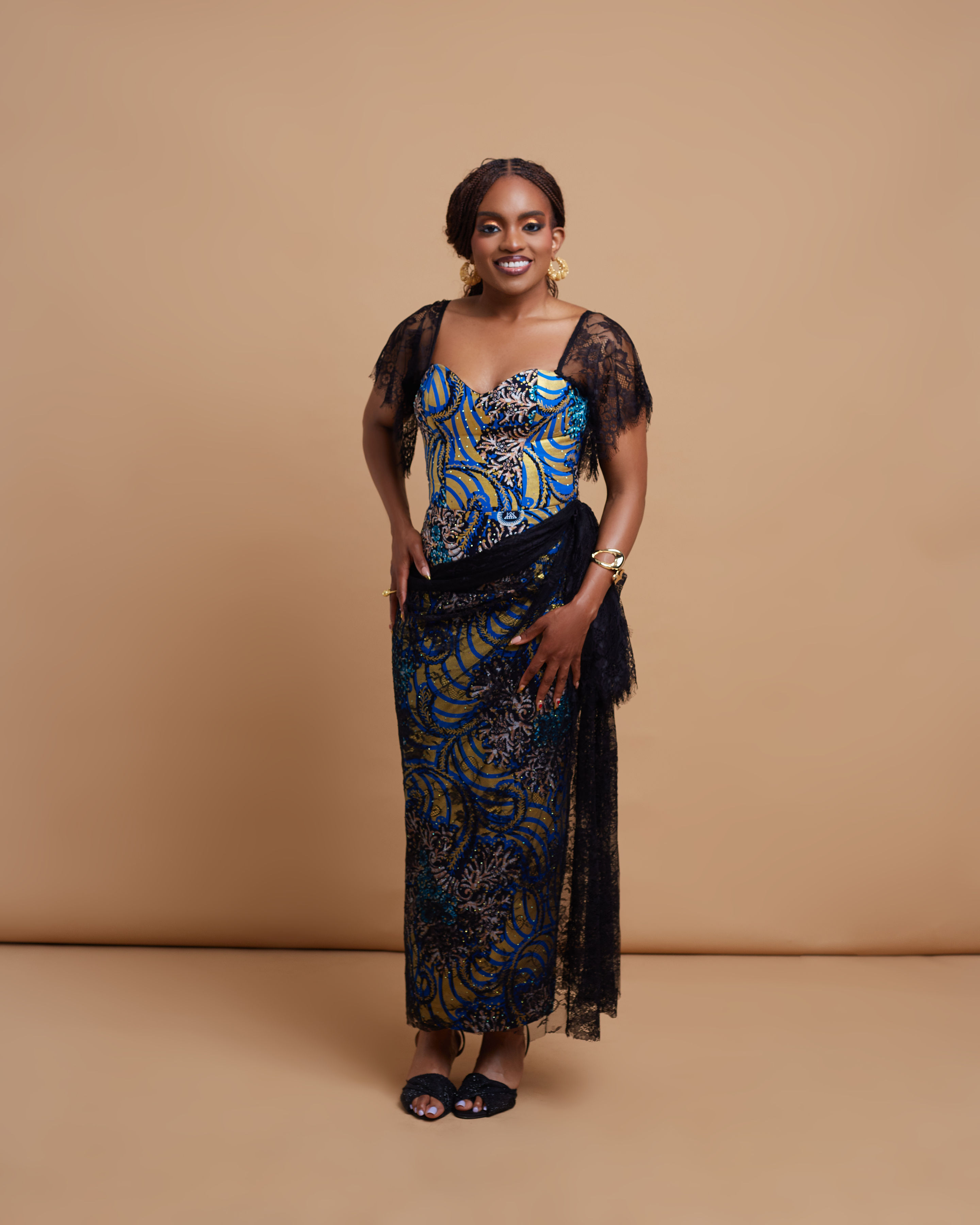 Half-length portrait of founder Adaobi Onyekaba in the Mmesoma — a blue and gold Swarovski stone wax Ankara corset gown with black Chantilly lace flutter sleeves and gold hoop earrings.