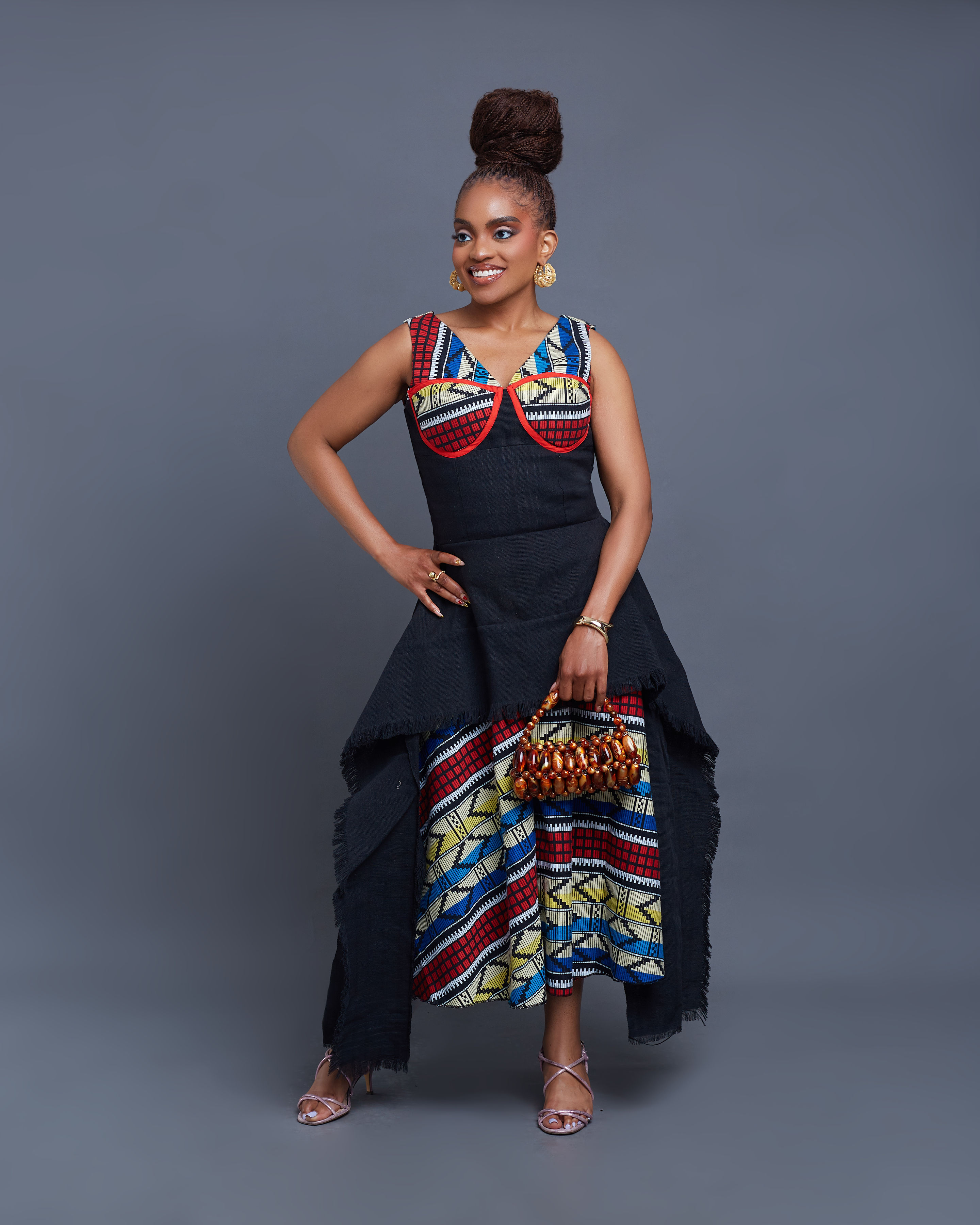Founder Adaobi Onyekaba seated with arms outstretched in the Ifeyinwa — a black Aso-Oke asymmetric dress with raw fringed hem over a red, cobalt, and white Akwete geometric panel — against a charcoal slatted wall.